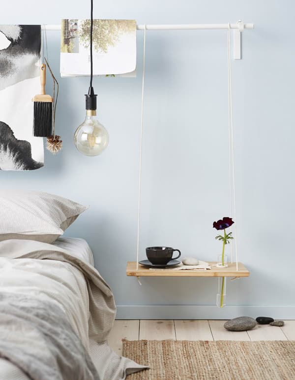 A DIY hanging bedside table made from a chopping board and rope has a hole drilled in it to hold a thin glass tube vase.