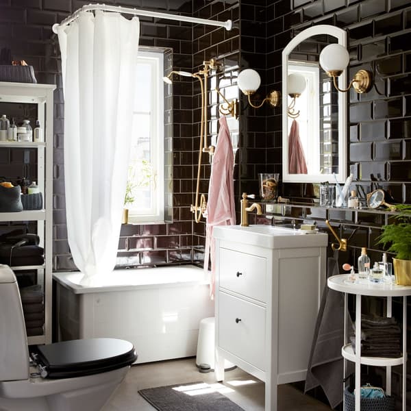 A dramatic and classic bathroom with glossy black tiles, white furniture, and brass-colored wall lamps by the mirror.