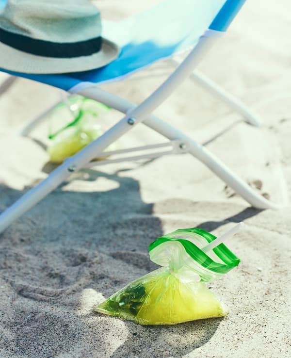 A drink served in a plastic bag sits in the sand next to a beach chair.