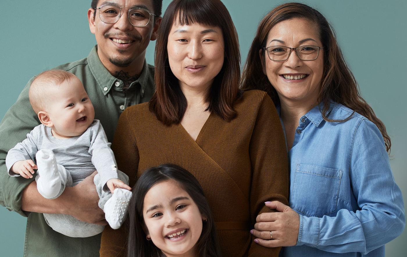 A family of five, all smiles from couple with a baby, a young daughter and a grandmother standing close together.