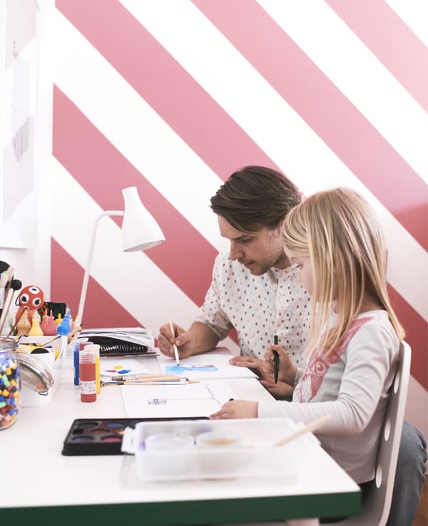 A father and daughter sat painting at a desk in front of brightly-coloured wallpaper.