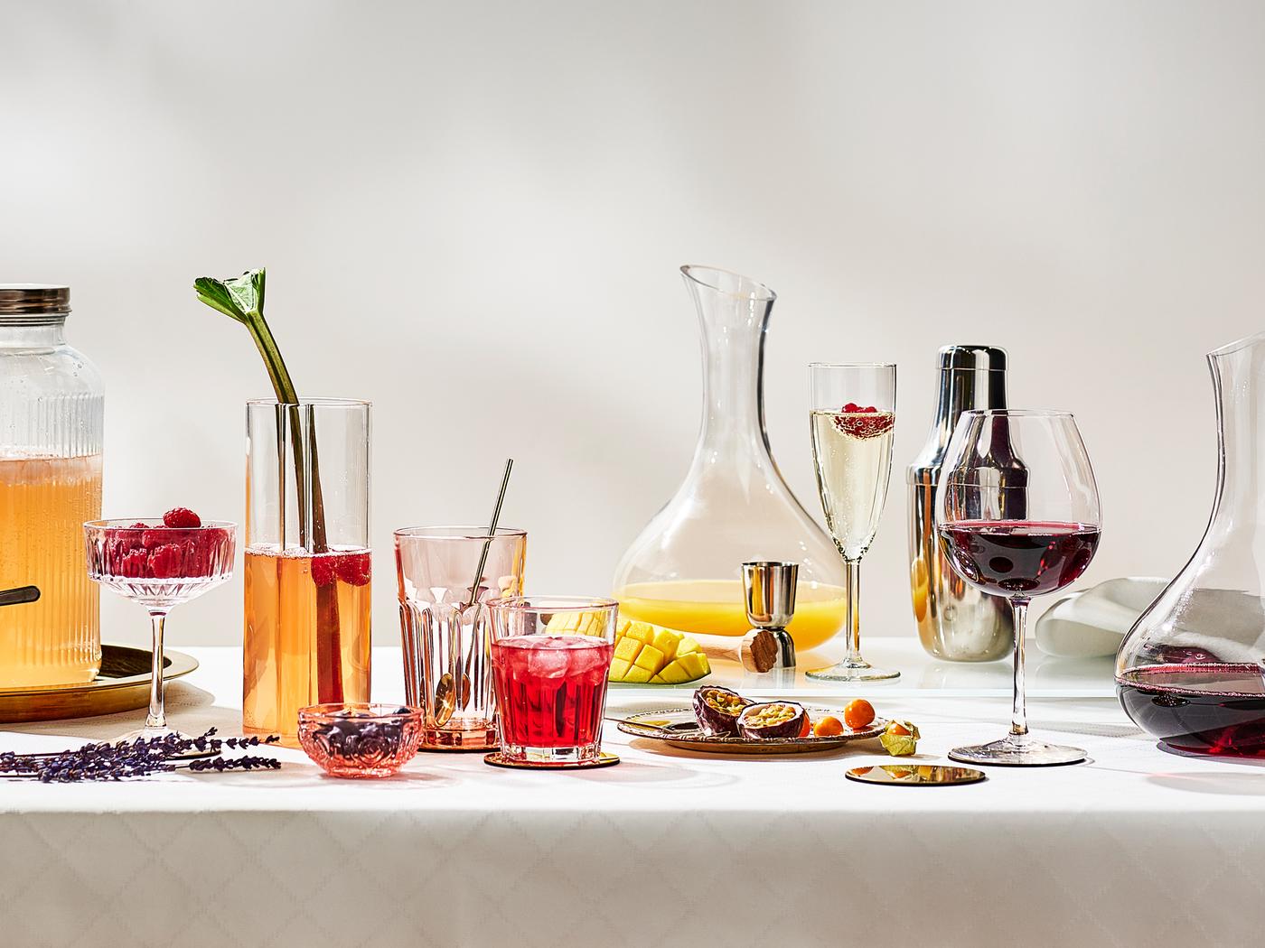 A festive table with fruits and various drinking glasses with different drinks in them on a white tablecloth.