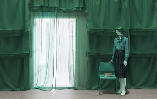 A figure dressed in green standing next to a green chair with a backdrop of green layered curtains.