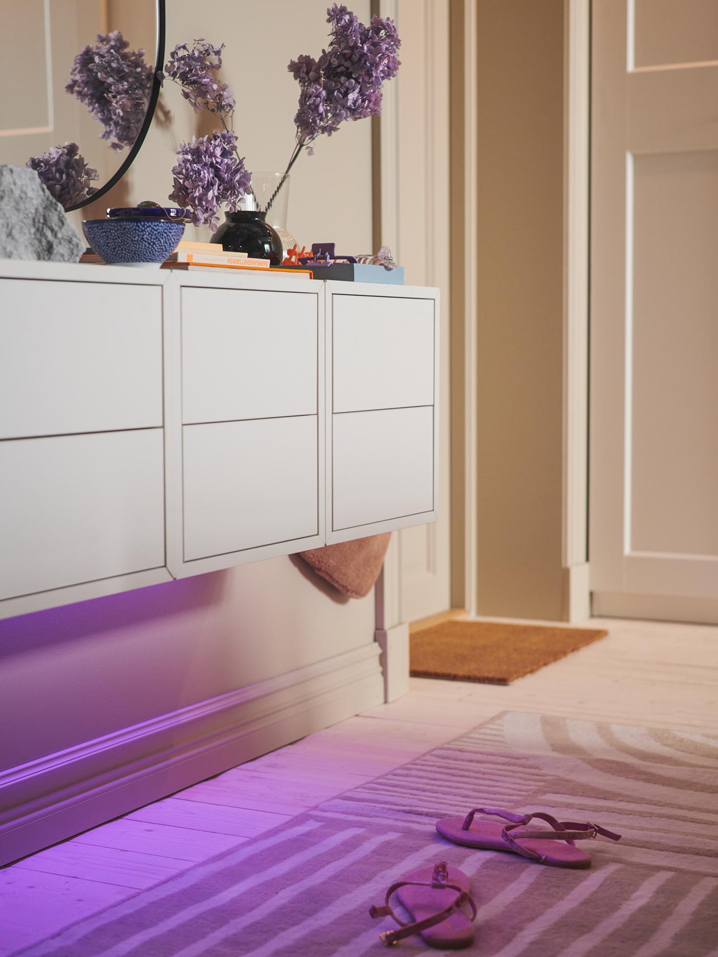 A floor is bathed in purple light by a LED lighting strip under three EKET cabinets with 2 drawers mounted on a wall.