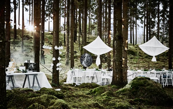 A forest wedding dining setup with a long white table, white chairs, decorative hanging objects and a cake table