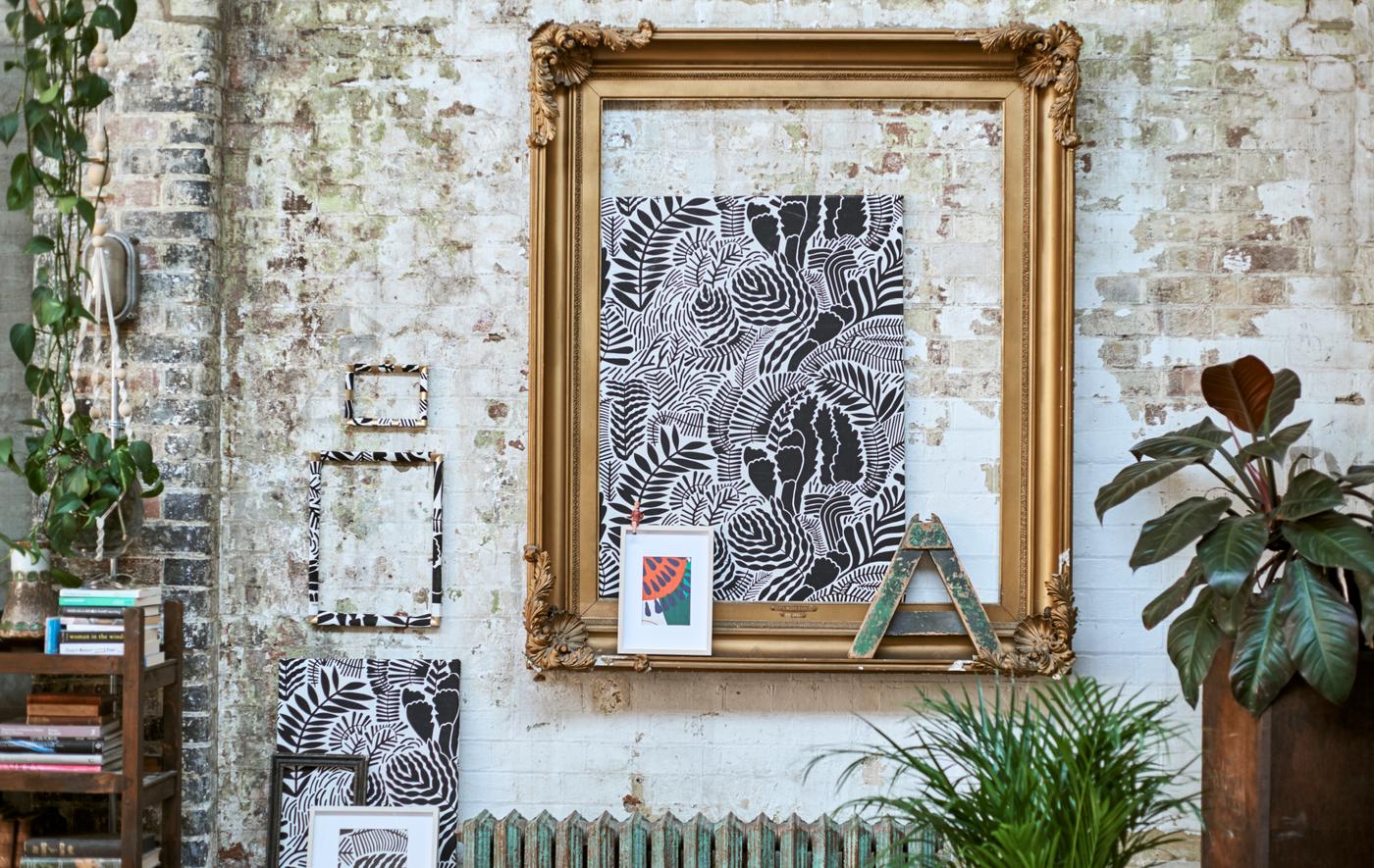 A gallery of framed patterned fabrics displayed on a brick wall with a large gold frame, hanging plants and a low bookcase.