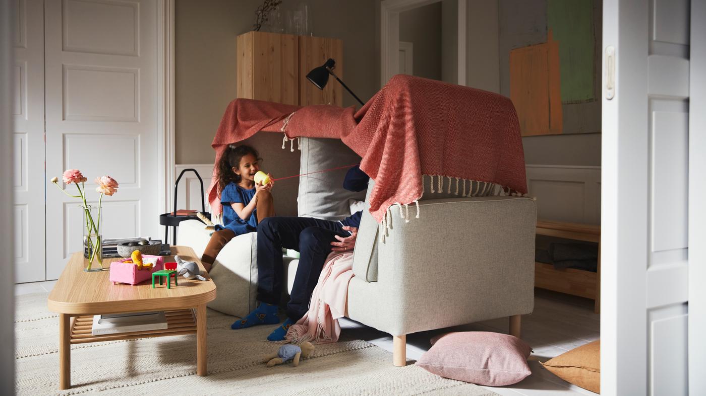 A girl and a man playing inside a blanket fort built on a beige PÄRUP sofa, beside an oak veneer coffee table on a beige rug.
