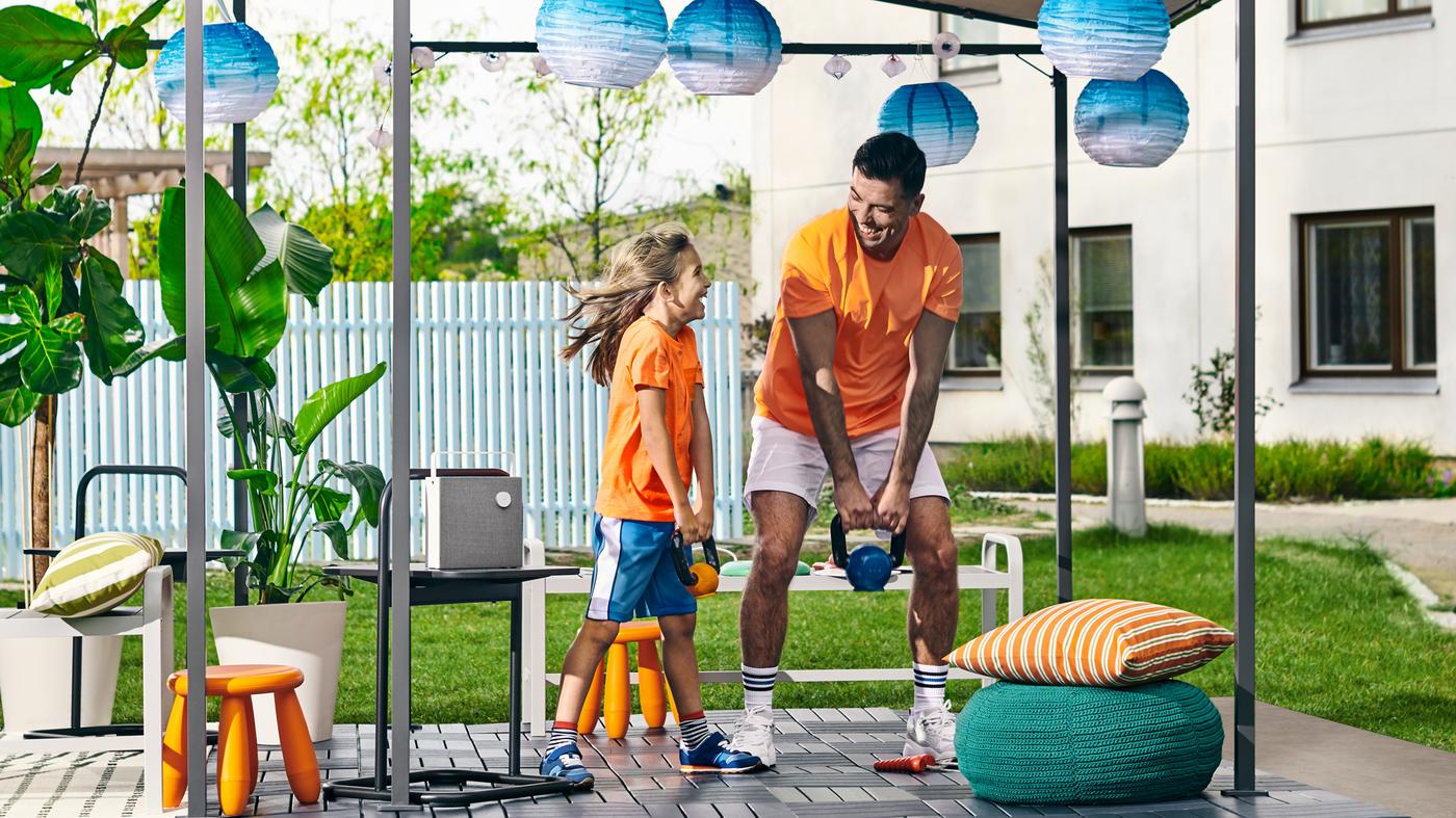 A girl and father lifting weights in a backyard with a GUNNÖN gazebo, RUNNEN outdoor floor decking and solar-powered lamps.