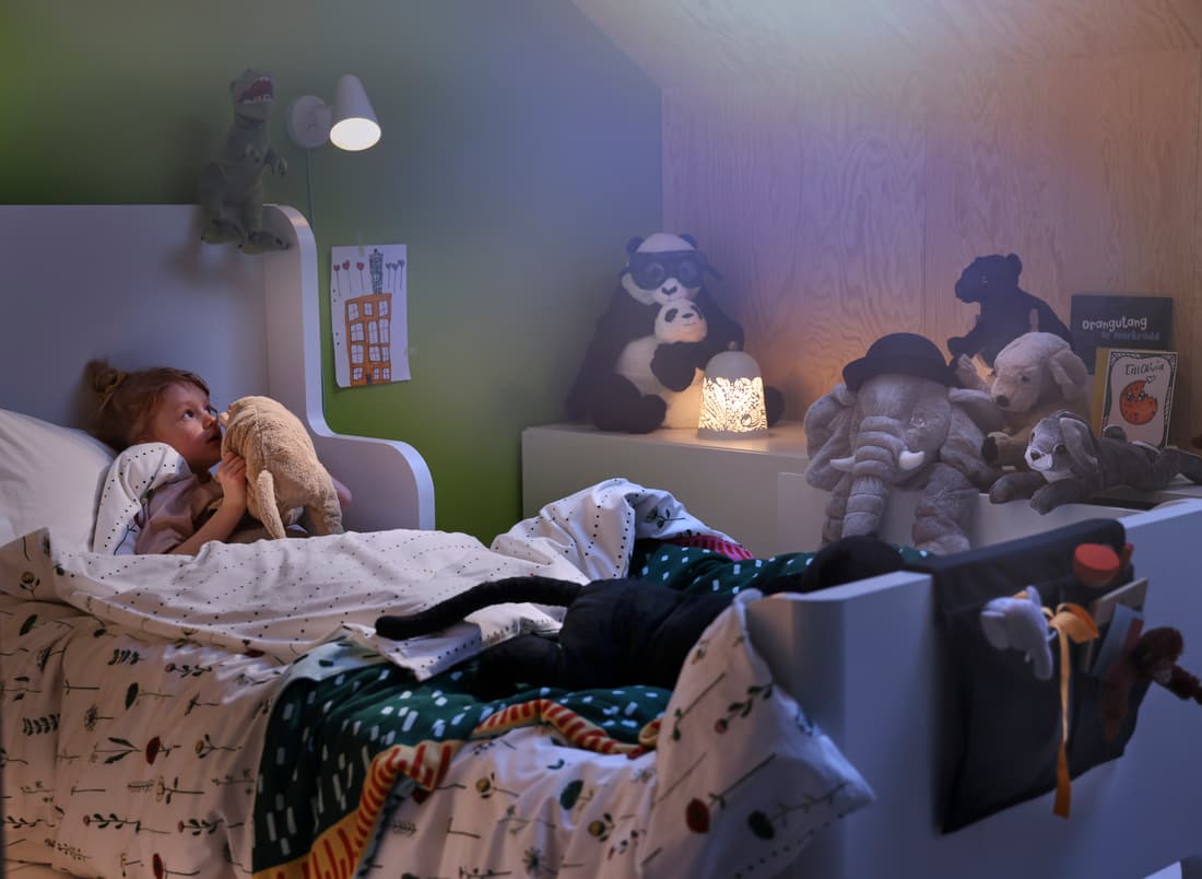 A girl cuddles her GOSIG GOLDEN soft toy dog in a BUSUNGE extendable bed with dim warm yellow lights. She is covered with a white KÄPPHÄST duvet with patterns of trees and is surrounded by animal soft toys.