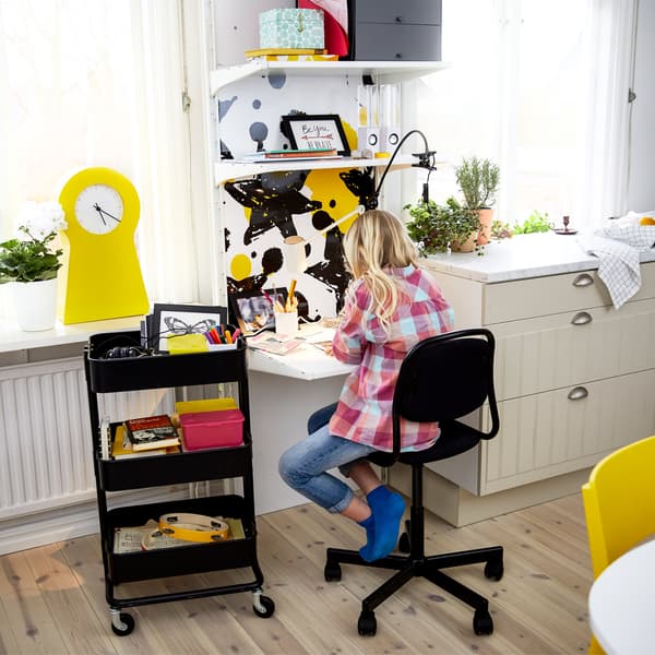 A girl doing her homework at a small desk top mounted on the wall and above the desk several shelves storing boxes.