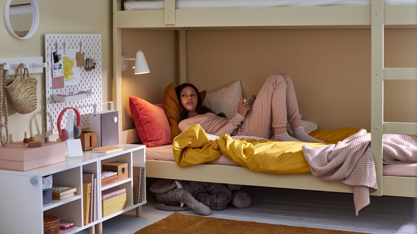 A girl in a bottom bunk bed with bed linen in mixed colors, a soft toy elephant under the bed and various white storage.