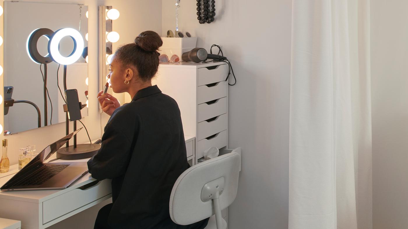 A girl is in her dorm on a BLECKBERGET swivel chair at a MICKE desk. She applies makeup and a LÅNESPELARE ring light is on.
