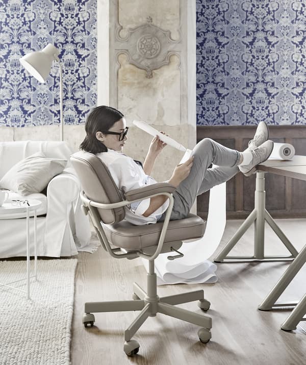 A girl leaning back in a beige swivel chair with her feet up on a desk in an office with blue patterned wallpaper.