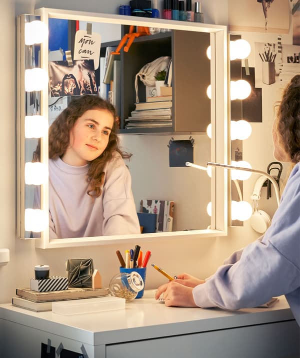 A girl looking into a mirror with lights above a white desk.