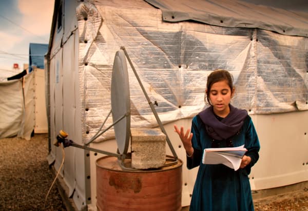A girl reading from a book in front of a makeshift satellite and temporary buildings.