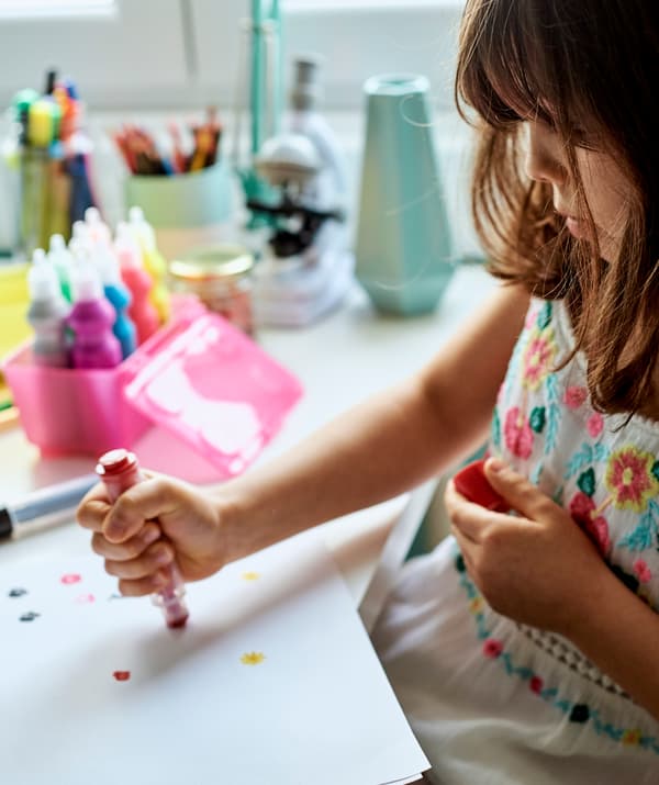 A girl sits at a desk using a red stamp pen to make patterns on paper, there are tubes of paints in a pink tub beside her.