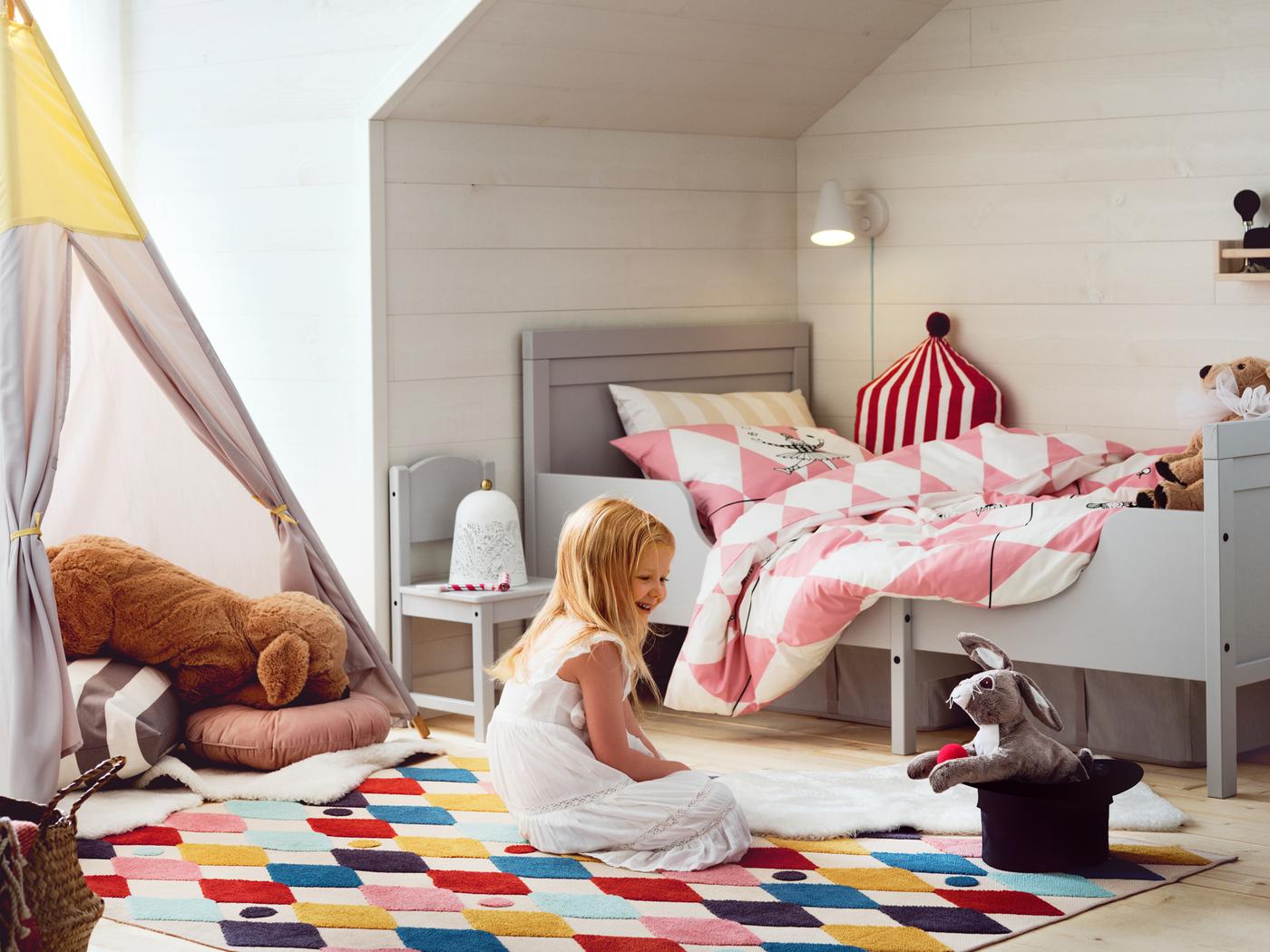 A girl sits on a brightly coloured BUSENKEL rug in a bedroom near a grey SUNDVIK extendable bed and looks at a toy animal.