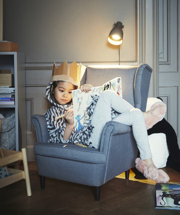 A girl wearing a cardboard crown, colouring while curled up on a grey chair.