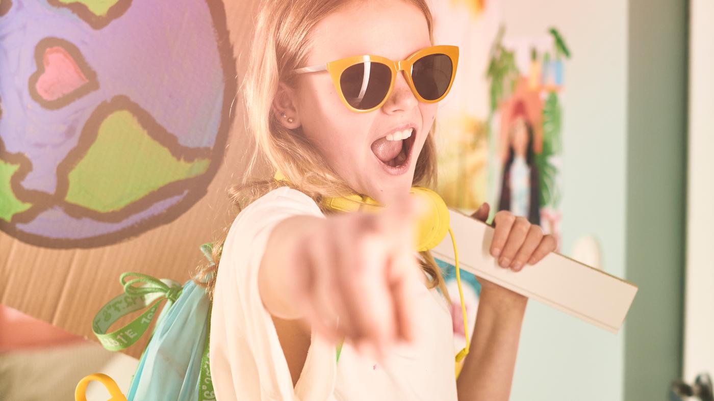 A girl, wearing yellow sunglasses, points forward, wearing a BLÅVINGAD blue-green backpack on her back.