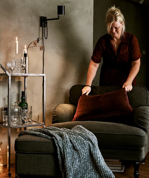 A green armchair next to a drinks trolley with cut glassware, at the chair a woman stands plumping a red velvet cushion.