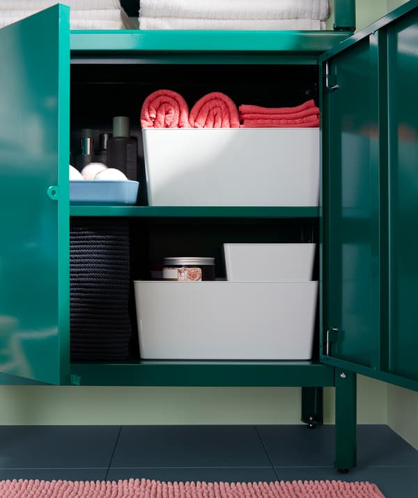A green metal cabinet, doors ajar, filled with various boxes and baskets, in turn filled with bathroom accessories.