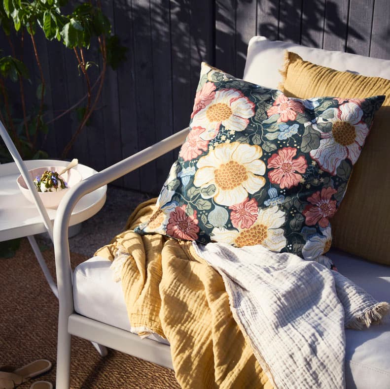 A green multicolour with floral patterned ARENBOK cushion cover is being used on a FJÄDRAR cushion pad placed on a sofa.