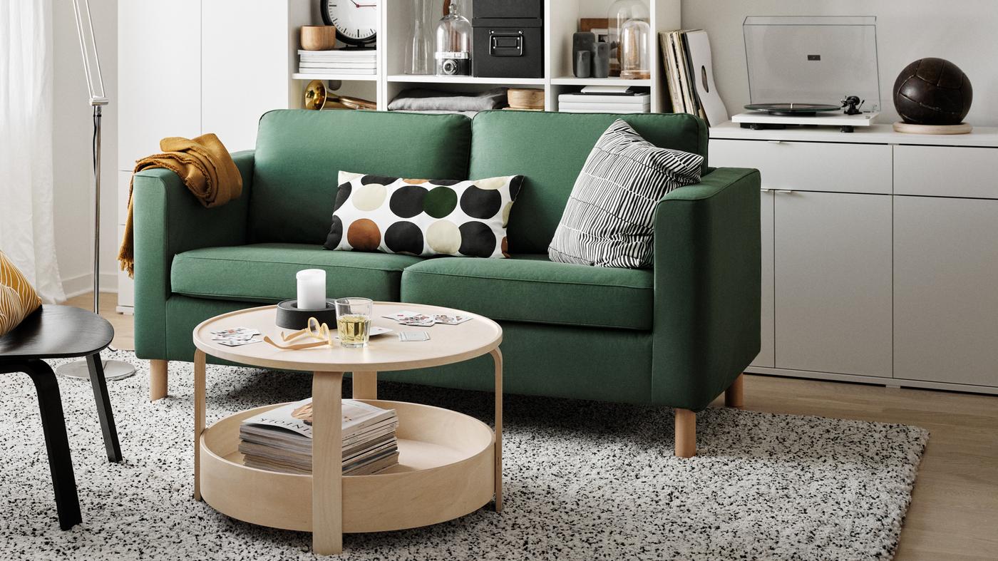 A green PÄRUP sofa with two patterned cushions on the seat in a white living room with open shelving behind it.