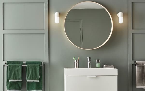 A green panelled bathroom with OSTANA wall lamps and a round mirror above a sink.