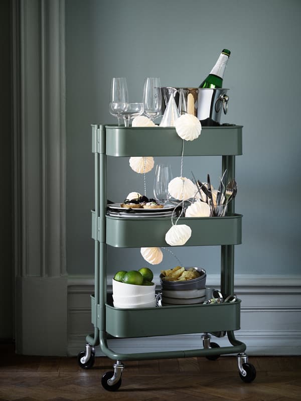 A green RÅSKOG trolley is packed with things for a party, including glasses, dinnerware and drinks to share.