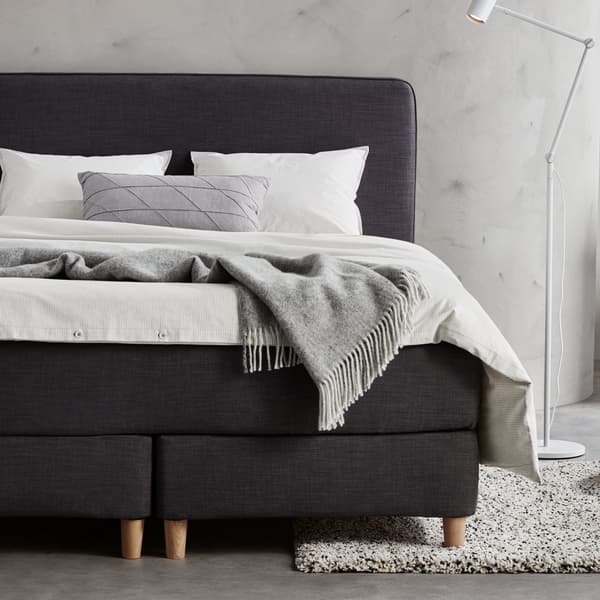 A grey bedroom with a dark grey DUNVIK divan bed covered with white bed linen. A white NYMÅNE floor/read lamp is beside it.