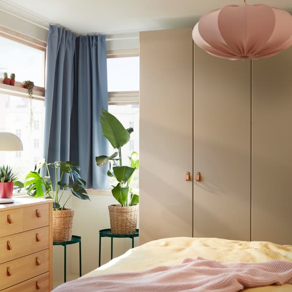 A grey-beige wardrobe, blue curtains, a pink pendant lamp, yellow bed textiles, a pale pink throw and two green tray tables.