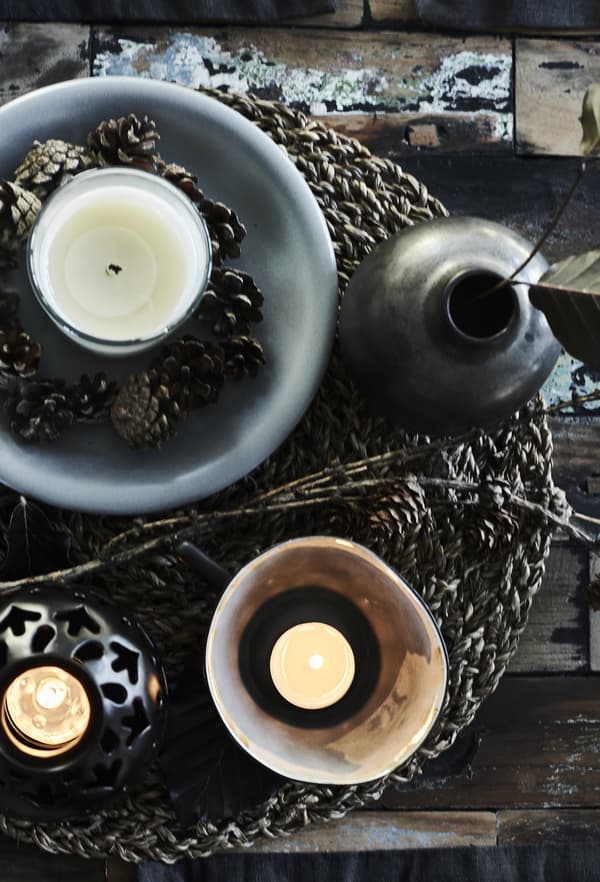 A grey bowl with a white candle in a glass and pine cones displayed on a woven placemat.
