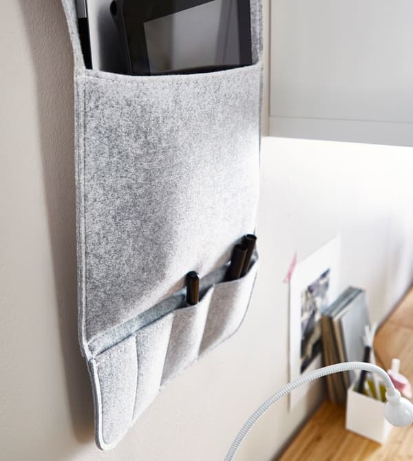 A grey fabric wall organiser hanging on a wall above a desk, with multiple pockets to hold pens and office supplies.