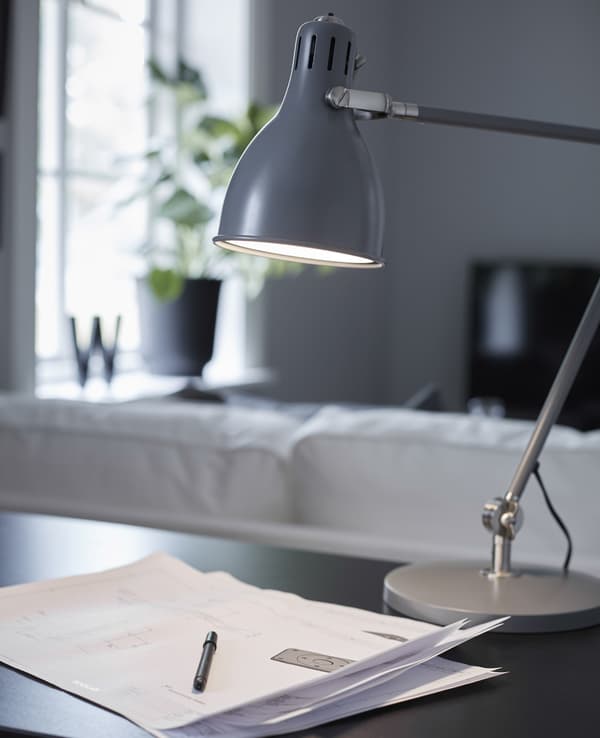 A grey metal office desk lamp shines light on some papers and a pen on a black desk.