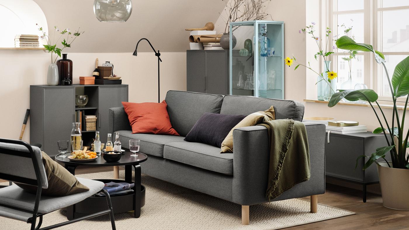 A grey PÄRUP sofa with three cushions on it, in a beige living room. A coffee table with drinks and snacks on top is in front.