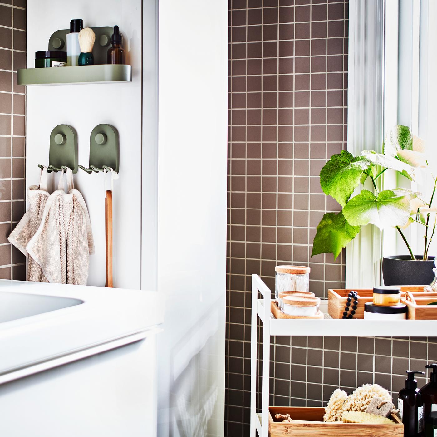 A grey-tiled bathroom with a white wash-stand, a white high cabinet, a white trolley with trays and jars plus a potted plant.
