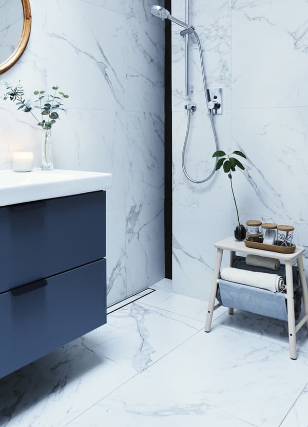 A grey washstand in a bathroom with white marble wall tiles, a wall-mounted shower and wooden stool with jars on top.