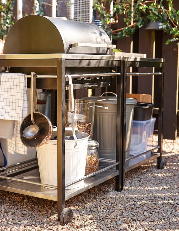 A grill with storage for charcoal or gas, sitting on gravel in an outdoor space.