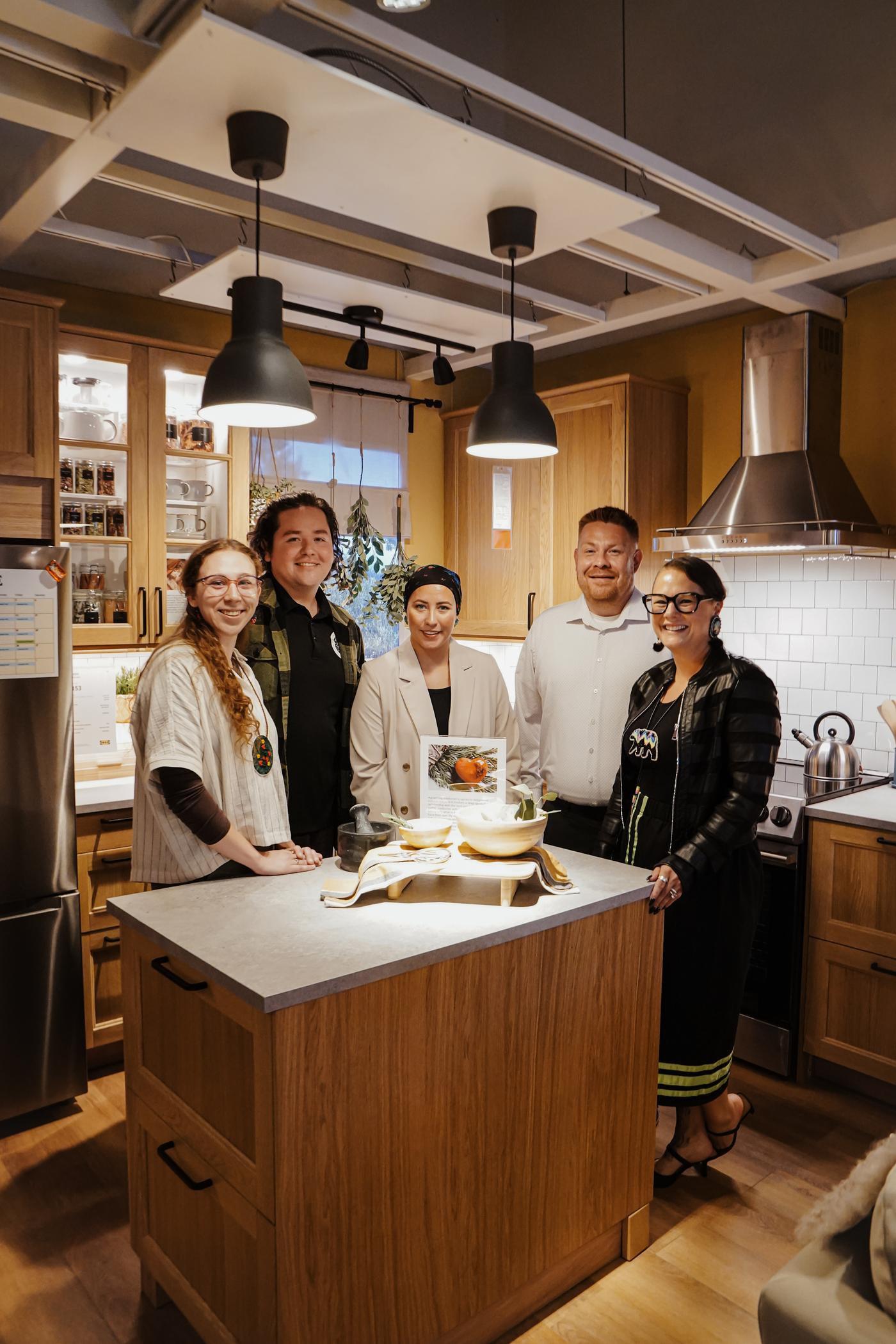 A group of people are gathered around a kitchen island and are looking towards the camera and smiling.