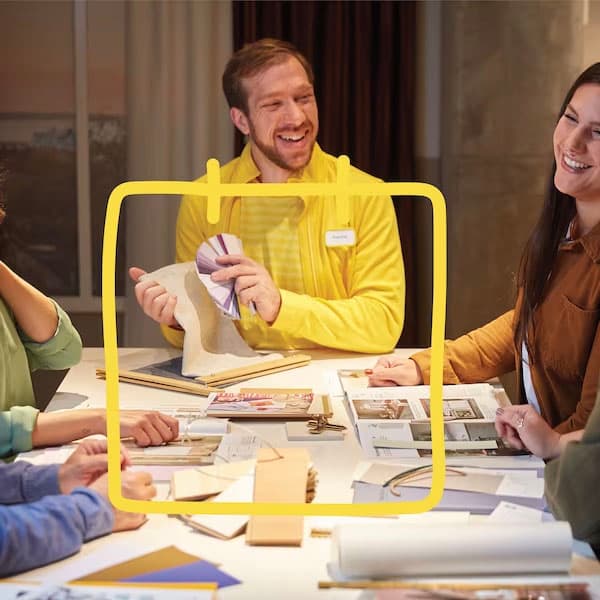 A group of people gathered around a table reviewing fabric samples, color swatches, and design materials, collaborating on a creative project in a well-lit workspace.