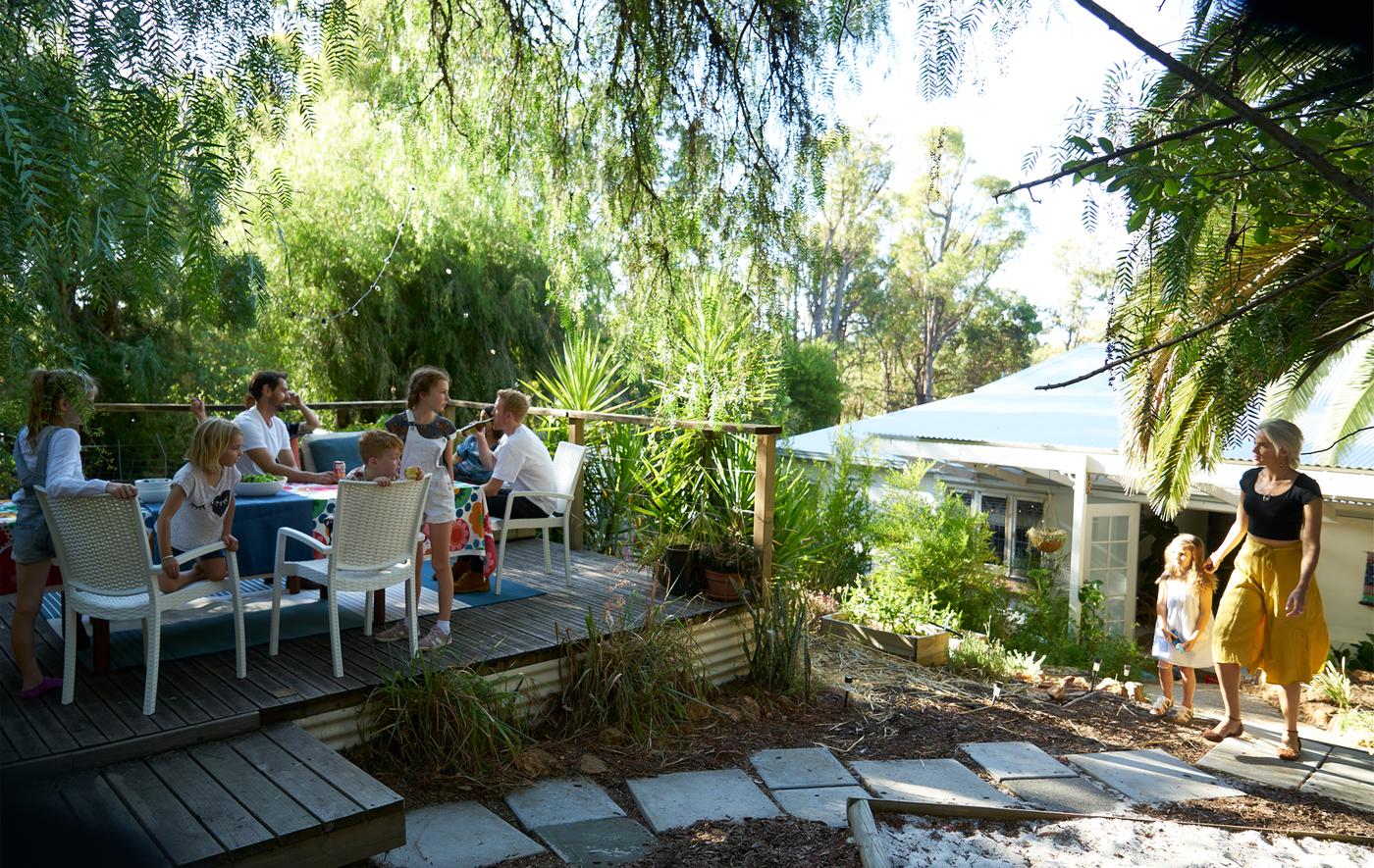 A group of people sit around a table on a raised deck in a garden with greenery and paving stones.