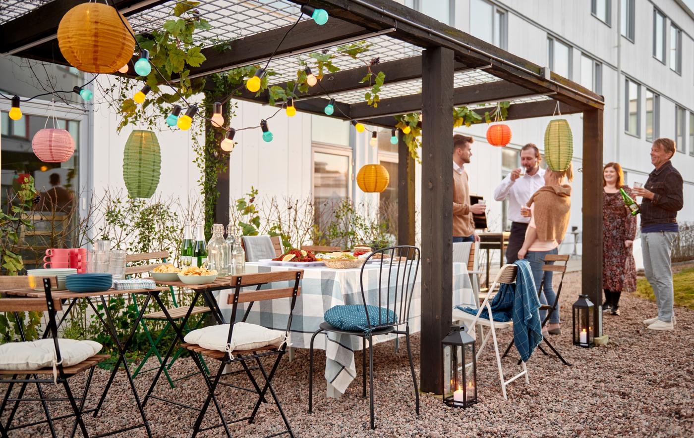 A group of people standing by a row of chairs and small tables set for a party under a decorated pergola in a courtyard.