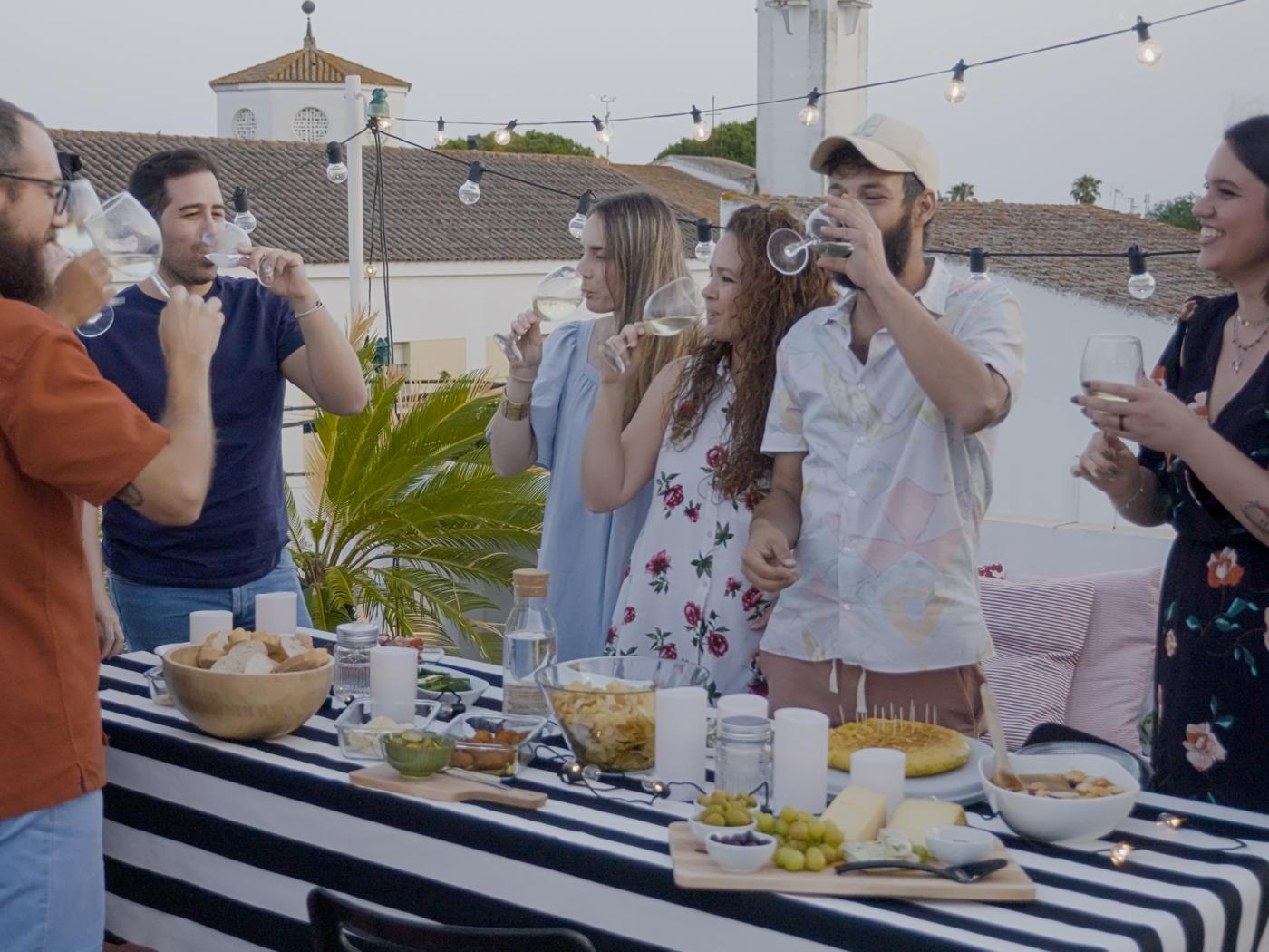 A group of young friends are cheerfully eating, drinking and talking, while standing at a buffet table full of food on an outdoor terrace.