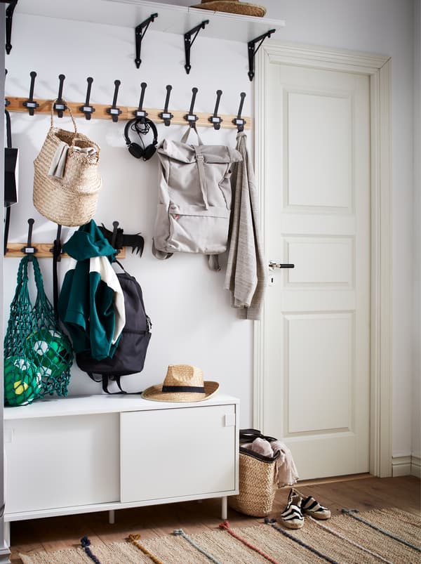 A hallway with hooks holding jackets and bags, a broad shelf high up and a white MACKAPÄR bench with storage compartments.