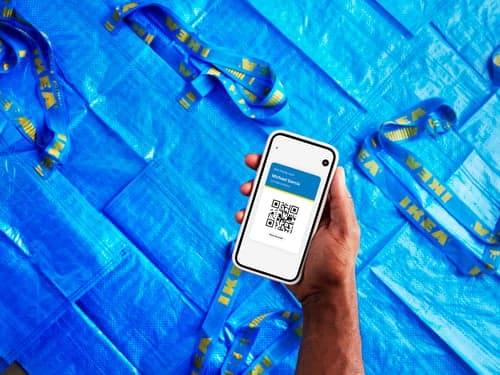 A hand holding a smartphone displaying the IKEA app above a background with several blue FRAKTA bags.