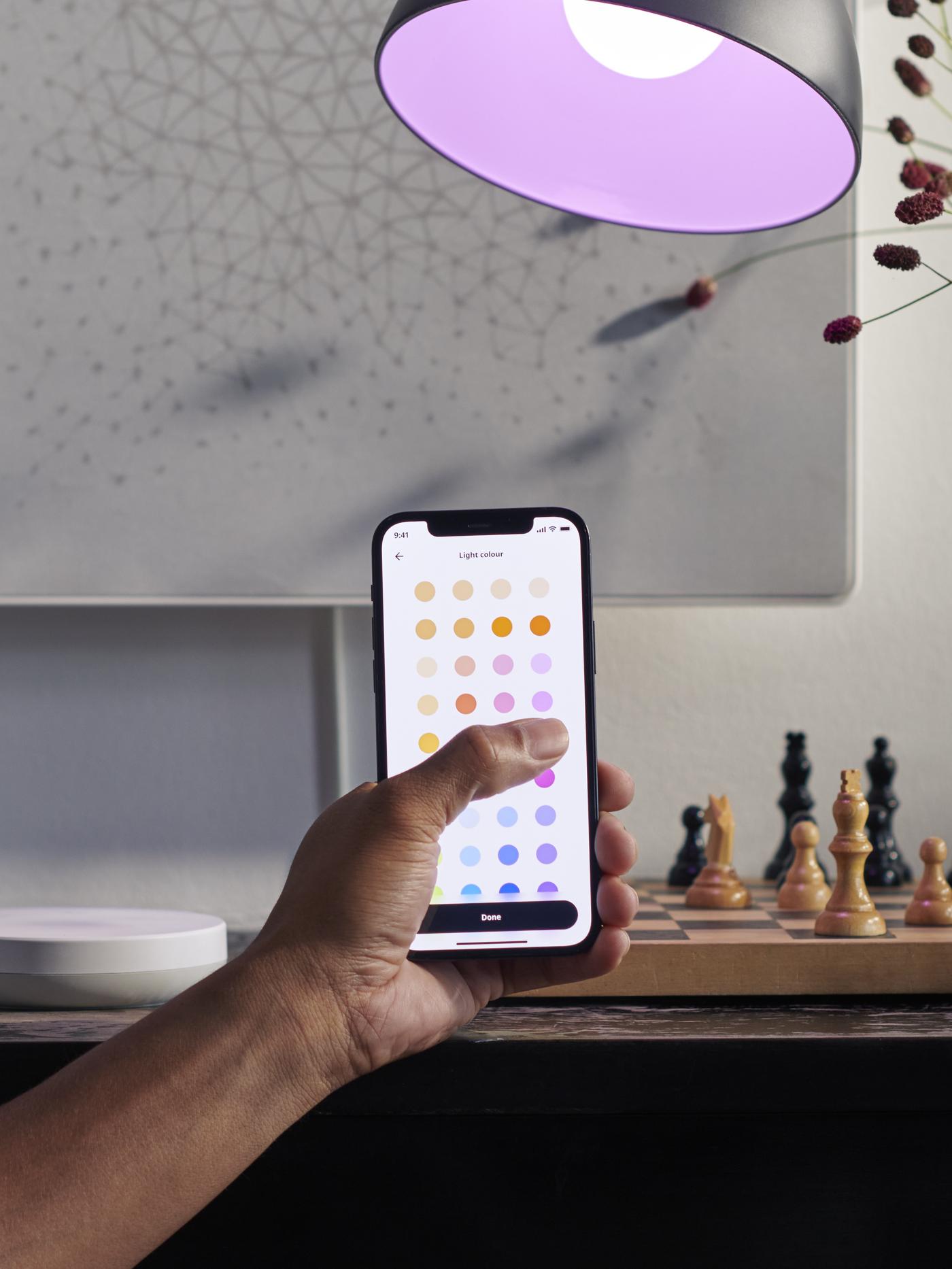 A hand holds a smartphone displaying color options, with a purple lamp overhead and chess pieces on a table.