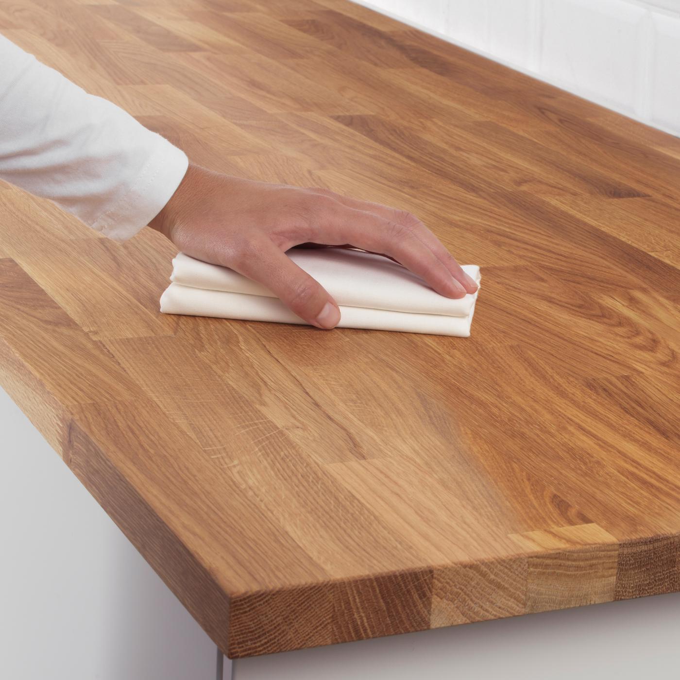 A hand holds a white cleaning cloth against the surface of a wooden kitchen worktop.