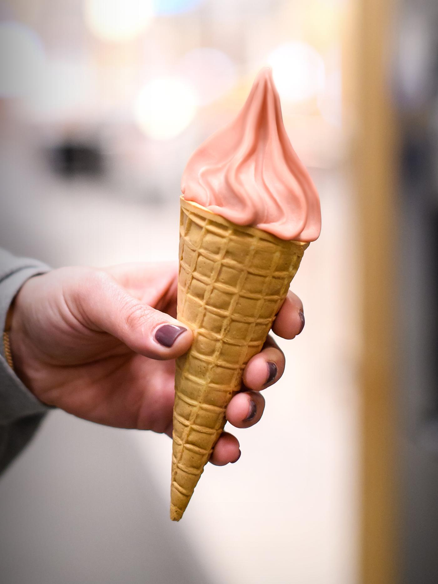A hand of a person wearing a grey top and purple nail varnish holding an ice-cream cone filled with strawberry soft ice.