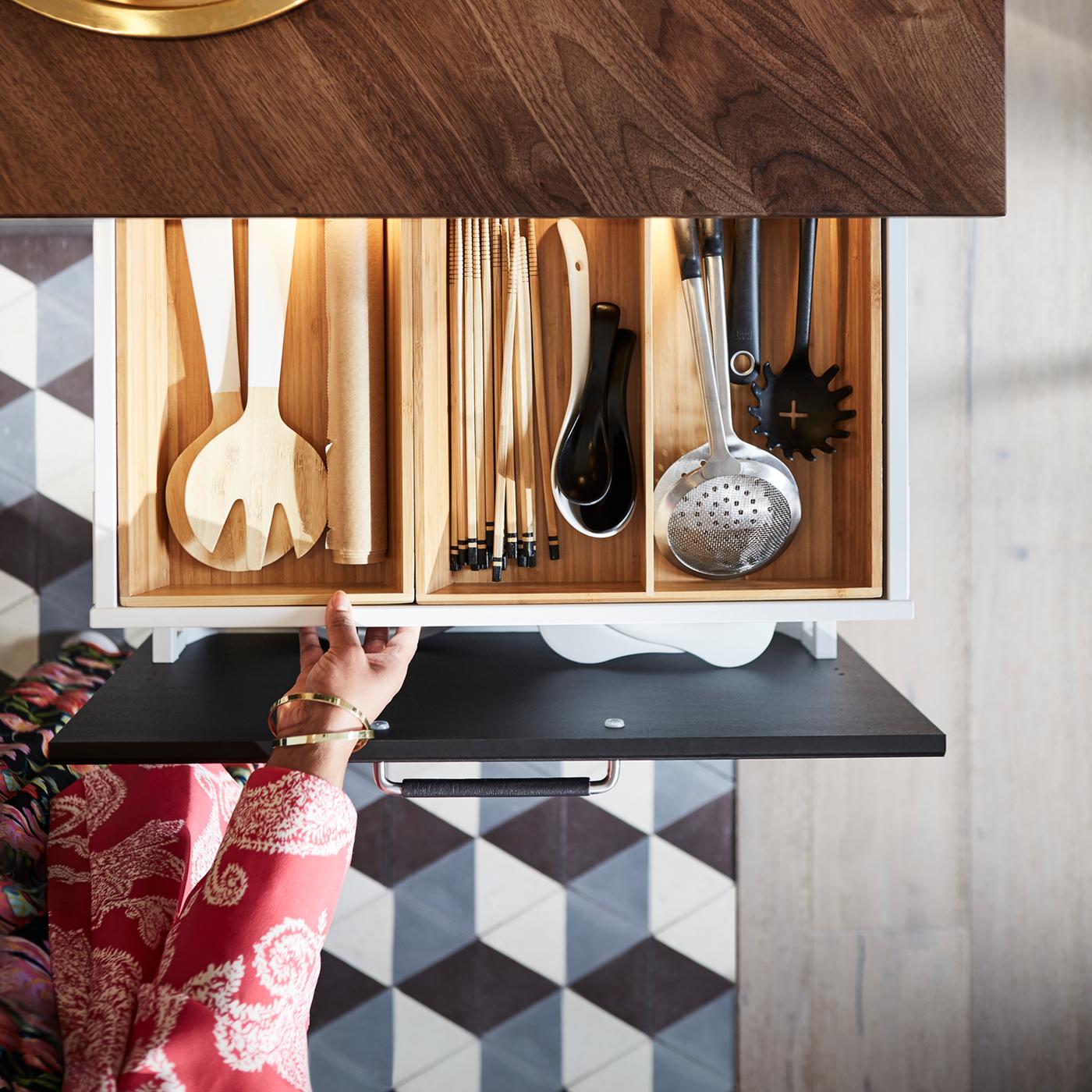A hand pulling out a drawer below a dark wood countertop with a wooden insert that’s filled with kitchen utensils.
