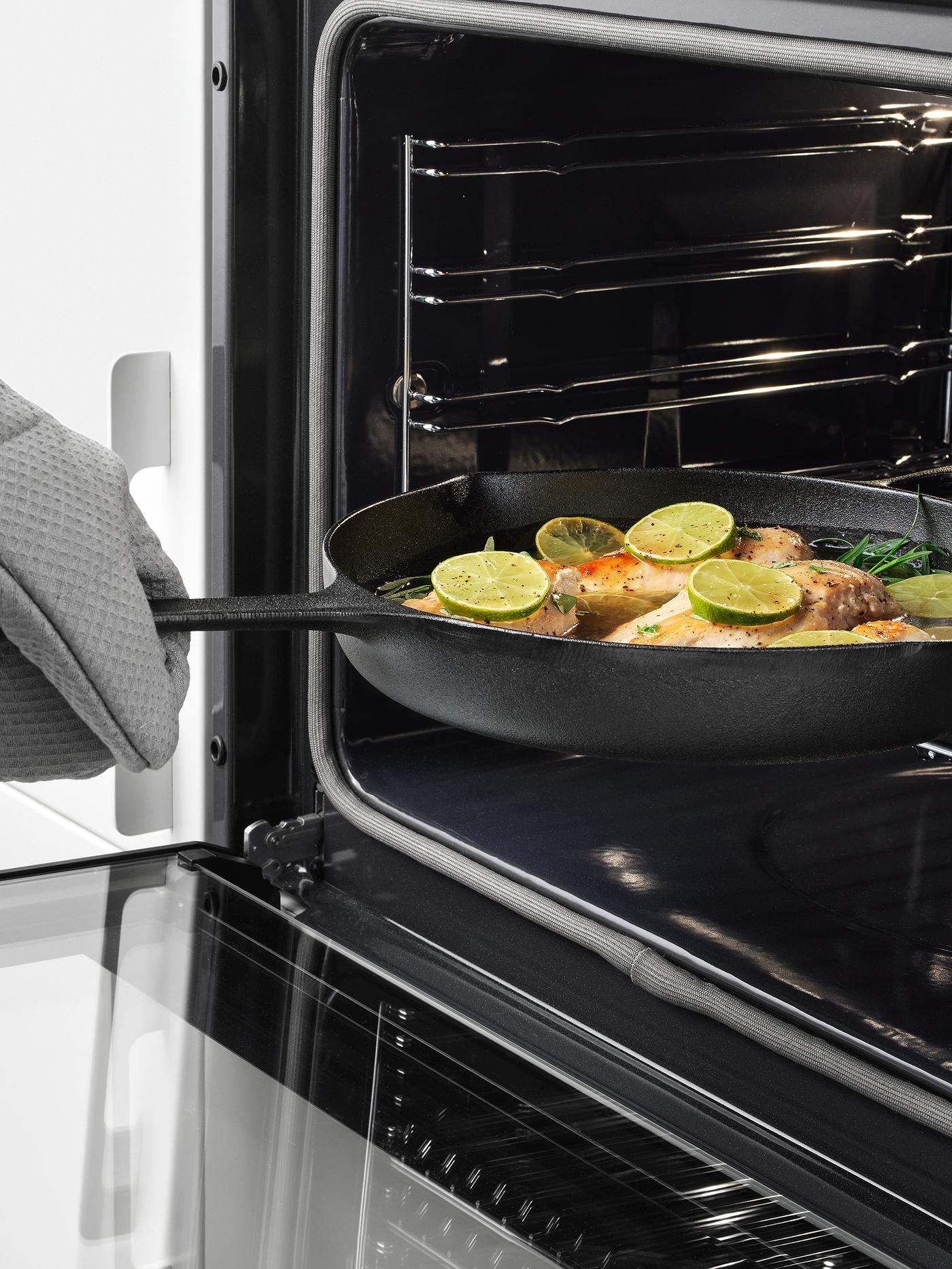 A hand wearing an oven glove placing a VARDAGEN frying pan in cast iron containing fish, lime and herbs into an oven.
